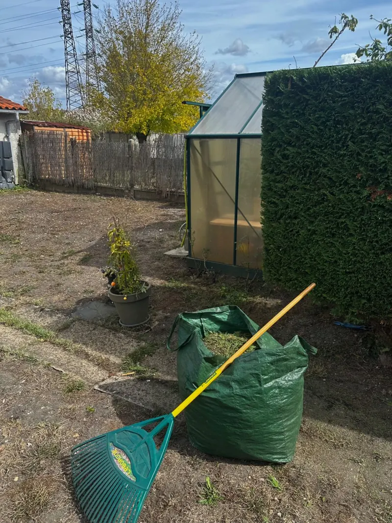 Sac de déchets verts et râteau devant une serre et une haie